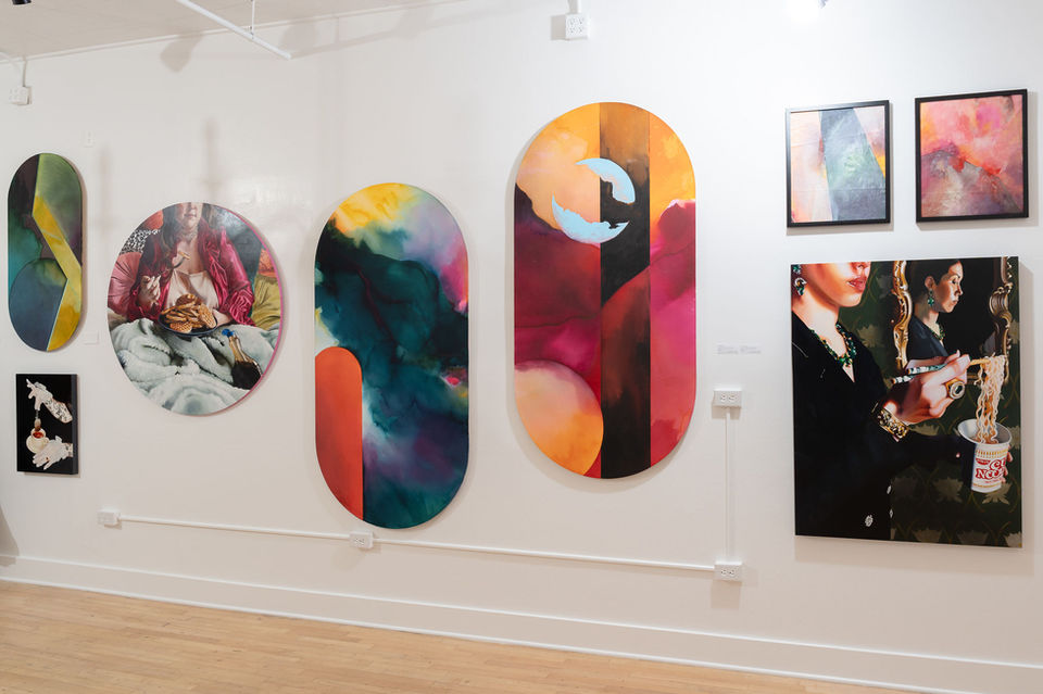 8 fine art paintings hanging on a white gallery wall. The paintings are colorful. 5 of the paintings are abstract, 3 of the paintings are representational. This is an art show put on in downtown Tacoma by Lydia Jewel Gerard and Amy Lewis.