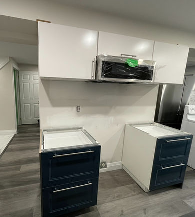 Two dark blue base ikea cabinets with drawers are positioned in the foreground, with ikea white upper cabinets and a stainless steel microwave covered in plastic wrap mounted above. The image captures a kitchen actively undergoing installation or renovation.
