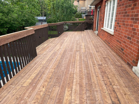 This image captures a newly pressure treated  deck extending from a red brick house, featuring a dark brown railing that curves around a view of a swimming pool. The spacious, rustic deck appears freshly prepared for outdoor enjoyment amidst a backdrop of lush green trees.