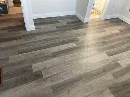A room is showcased with a newly installed grey wood-look vinyl plank flooring, creating a contemporary aesthetic. The neutral walls and white baseboards complement the floor, suggesting a freshly finished interior space.