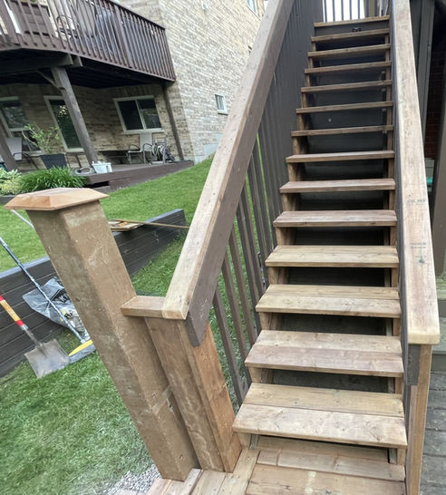 A sturdy pressure treated wooden staircase with a brown railing ascends towards an elevated deck attached to a brick house. The surrounding backyard area features a green lawn.