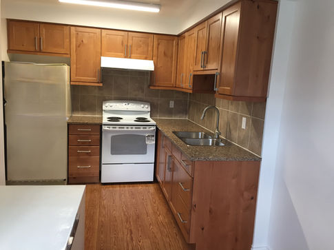 This kitchen features a stainless steel refrigerator, white stove, and a double sink set against light wood cabinetry and granite countertops. A tiled backsplash and warm wooden flooring contribute to the clean and functional appearance of the space.