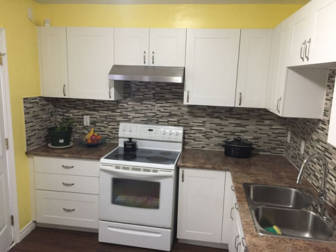 The central subjects of this image are the kitchen's white cabinets and stove, arranged around a striking mosaic tile backsplash and brown laminate countertops. A bowl of colorful fruit and a black pot are also visible on the counter, suggesting a functional and ready-to-use space.