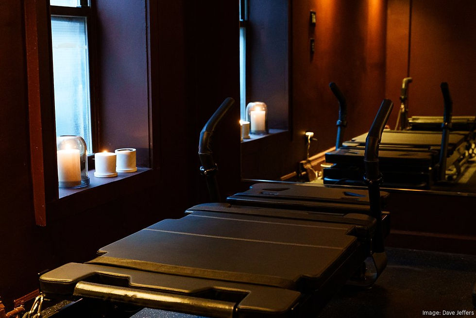 Lit candles on a windowsill create a serene ambiance in a dimly lit gym. A Pilates reformer is prominent against deep brown walls.