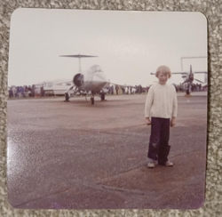 Photo of a young white boy with blonde hair standing on an airfield with a small military jet plane 
