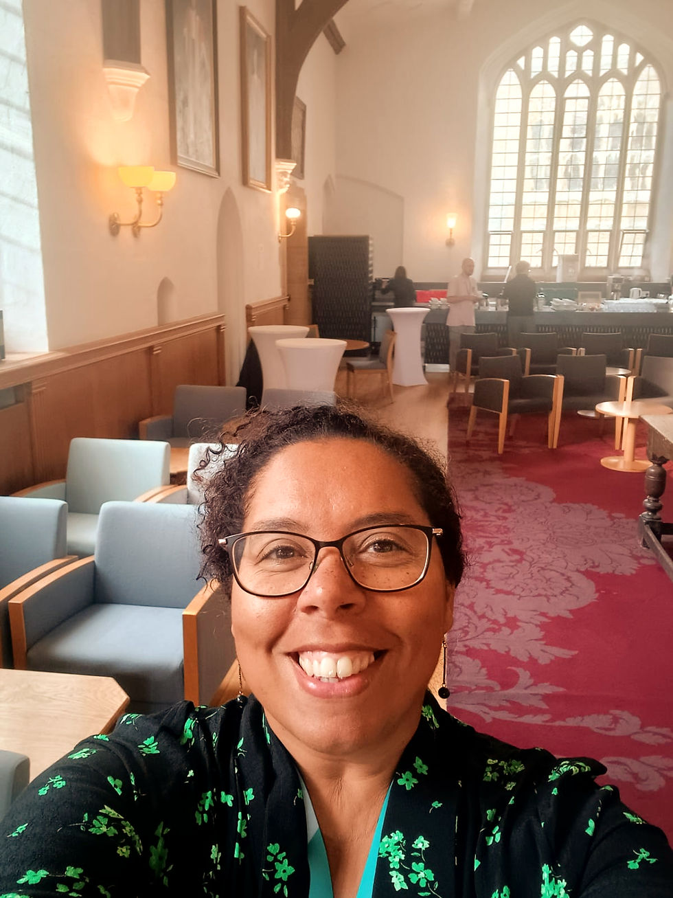 Smiling Black mixed race woman takes a selfie in a bright room with arched windows, elegant furniture, and red patterned carpet. Others chat in the background.