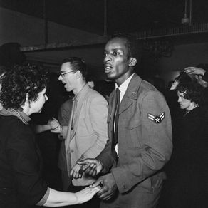 Black-and-white photo of a Black man in a 1950s USAF uniform dancing with a white woman on a crowded dance floor event, his face is intent and serious.