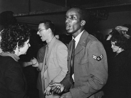Black-and-white photo of a Black man in a 1950s USAF uniform dancing with a white woman on a crowded dance floor event, his face is intent and serious.