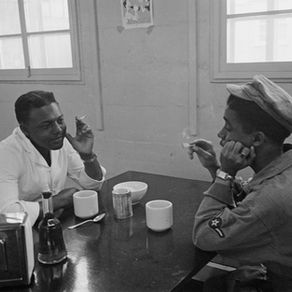 Black and white photo of two young Black men sitting in a canteen, with cups and condiments on the table. They're both smoking cigarettes. One is in a 1950s US Air Force uniform, the other is wearing a white shirt.