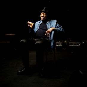 Seated older Black man in a hat and blue jacket holds a drink in a dimly lit setting. The mood is contemplative and relaxed.