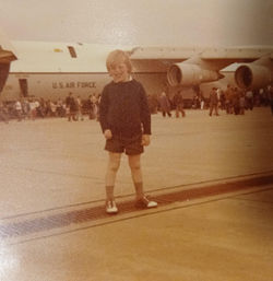 Photo of a young white boy with blonde hair standing on an airfield with a huge military jet plane i