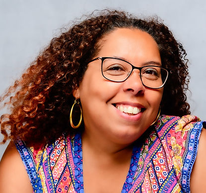 Black woman with shoulder length curly hair, glasses and hoop earrings smiles into the camera. She's wearing a colourful top.