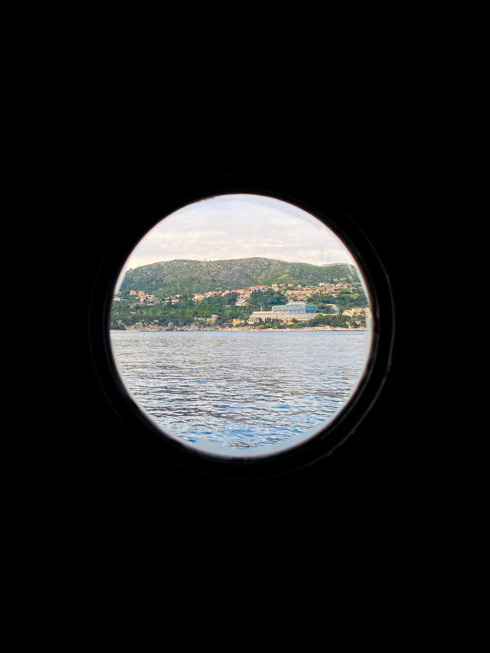 circular port window of a boat looking out at Mediterranean sea with green hills and stone houses in the background — travel photography by Shutter & Sojourn