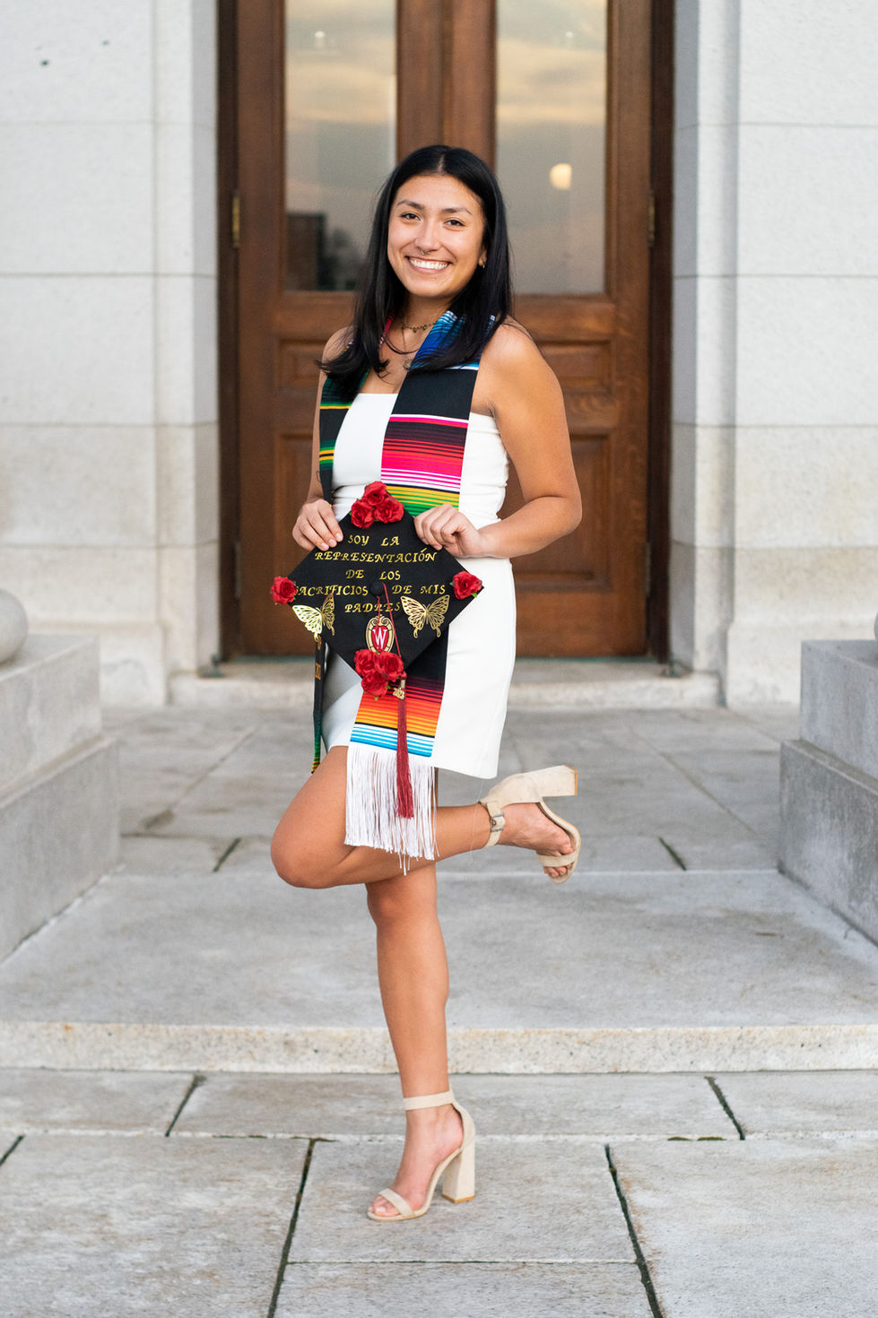Latina college graduate holding decorated cap with leg up at the WI state capital High school senior boy looking off into the distance in green field — graduation photography by Shutter & Sojourn