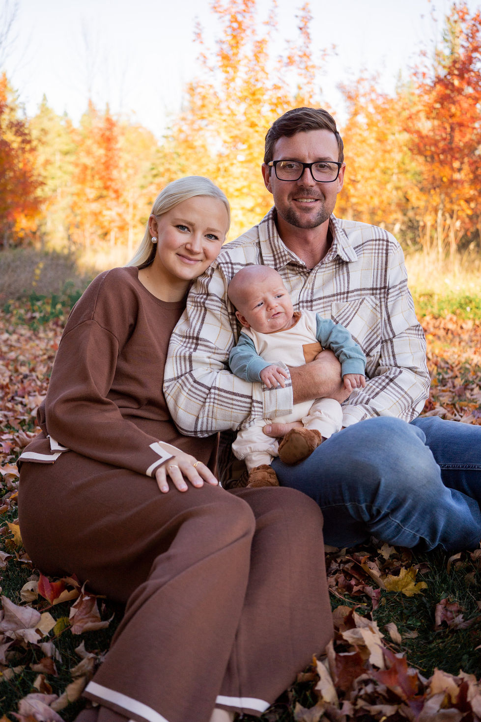 couple sitting on fallen leaves and holding baby while smiling at the camera during peak fall colors — family photography by Shutter & Sojourn