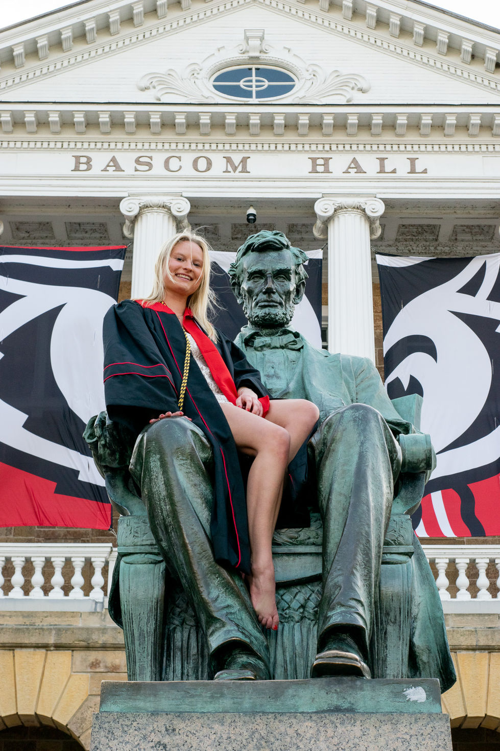 college graduate sitting on lap of statue at famous university landmark High school senior boy looking off into the distance in green field — graduation photography by Shutter & Sojourn