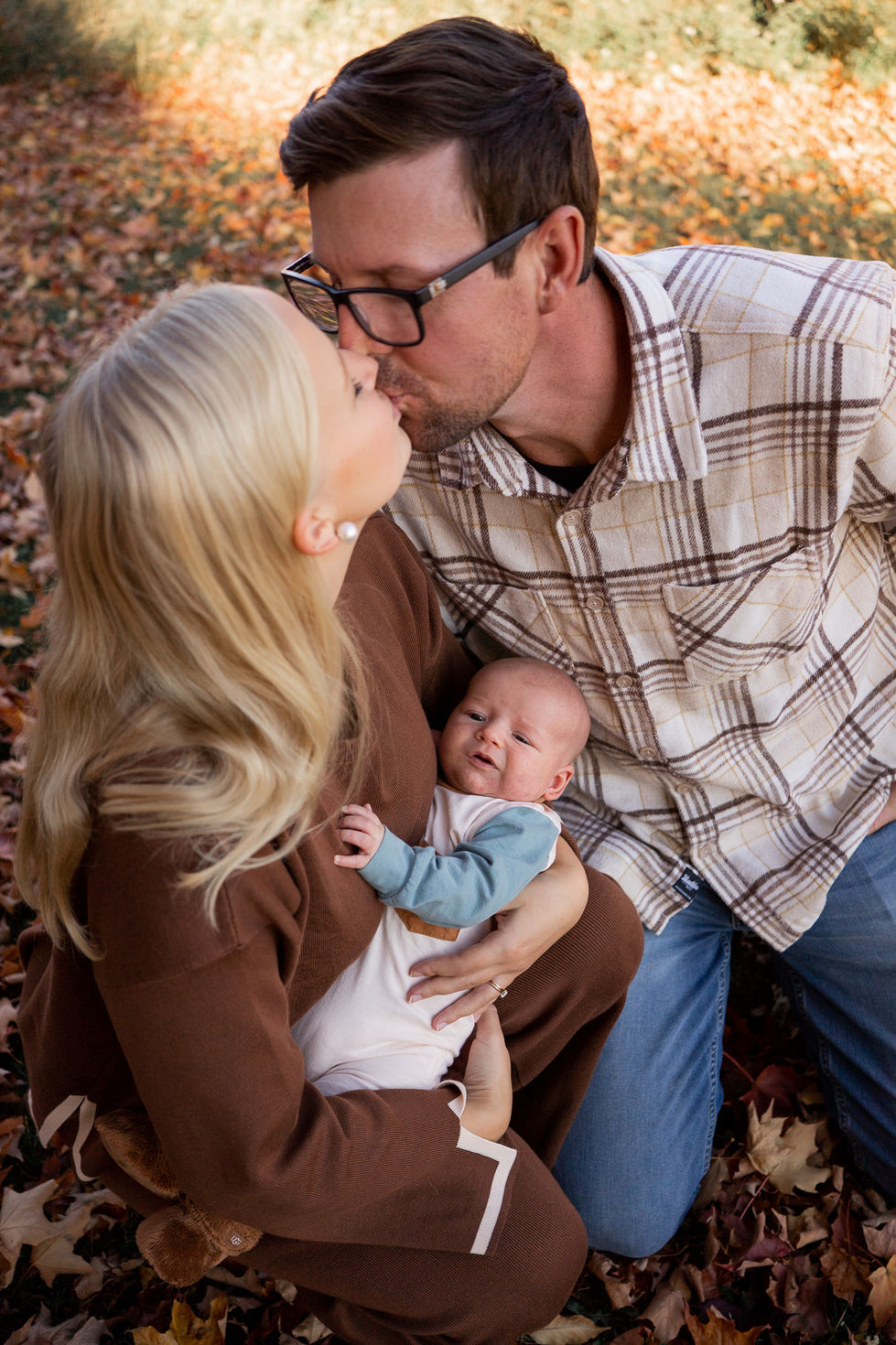 couple kissing while holding baby in blue onesie outside fall photo — family photography by Shutter & Sojourn