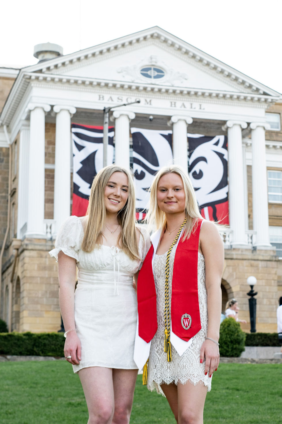 2 college graduates in white dresses and red ribbons smiling in front of university building