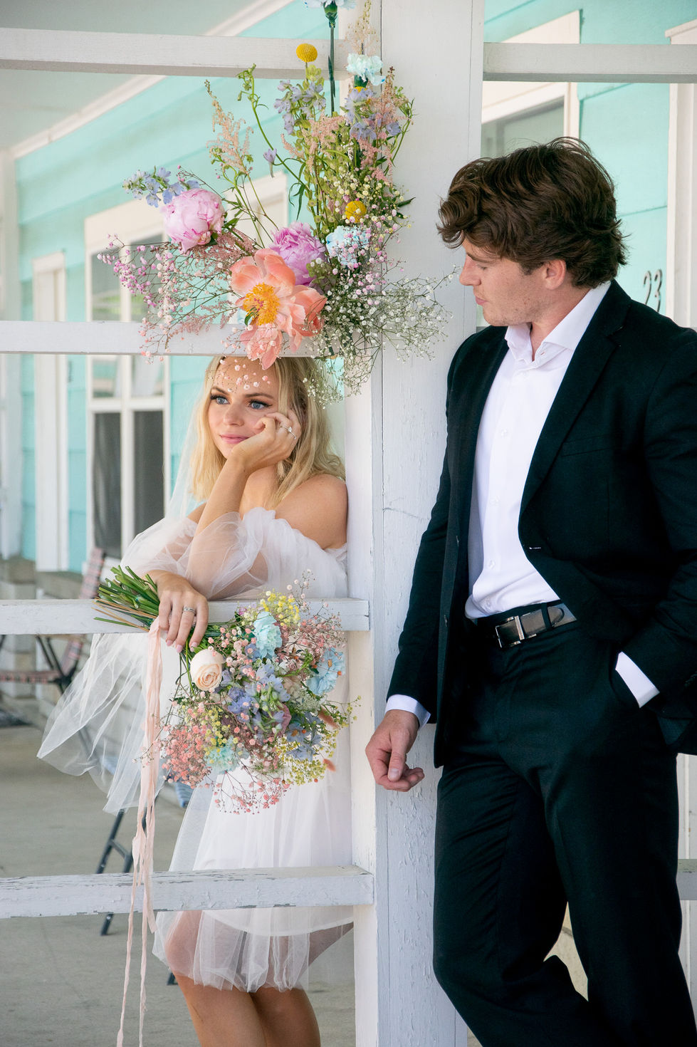 bride in short dress holding flowers and leaning on wall looking into distance while groom gazes at bride — wedding photography by Shutter & Sojourn