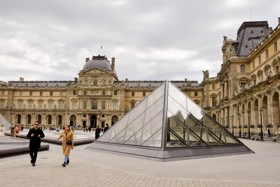 man dressed in black and woman dressed in tan walking in courtyard of the Louvre next to glass triangles — travel photography by Shutter & Sojourn