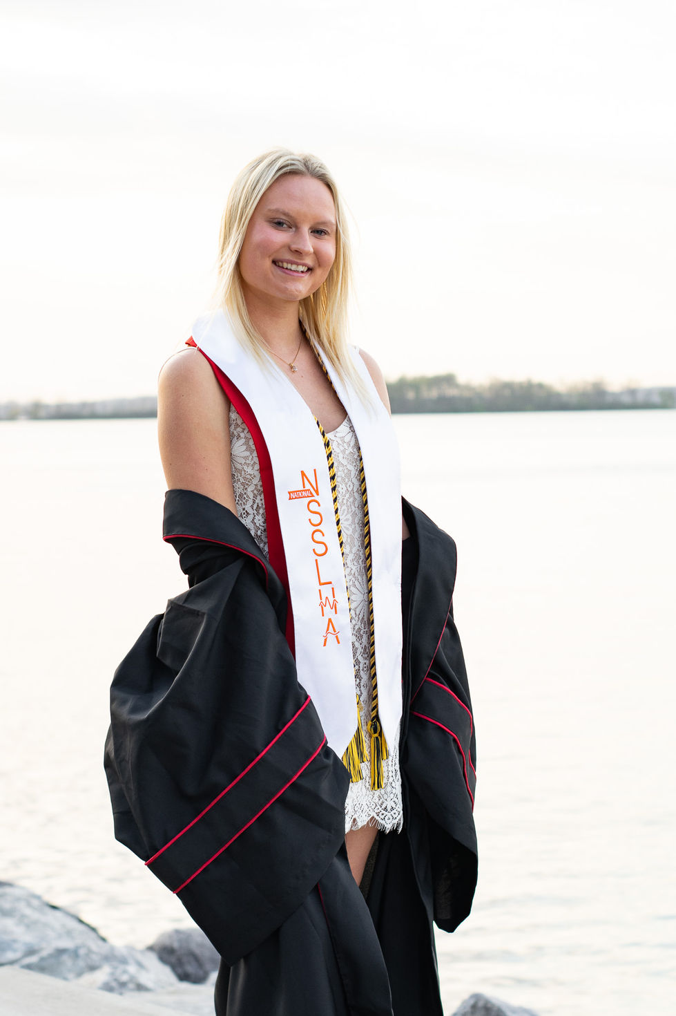 college graduate smiling in white dress with gown hanging off her shoulders on the waterfront High school senior boy looking off into the distance in green field — graduation photography by Shutter & Sojourn