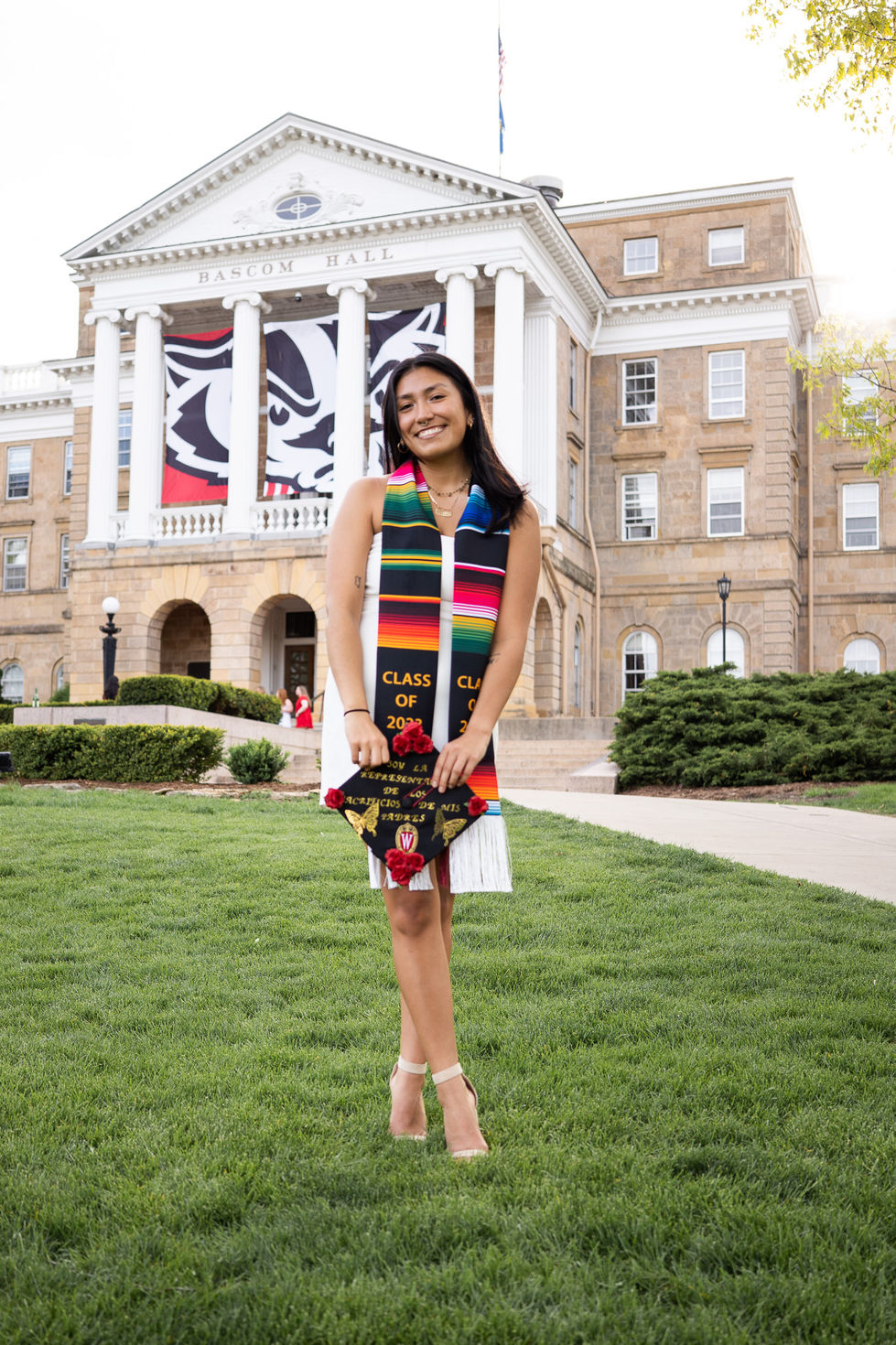latina college graduate in white dress and ribbon holding cap in both hands with legs crossed on Bascom hill High school senior boy looking off into the distance in green field — graduation photography by Shutter & Sojourn