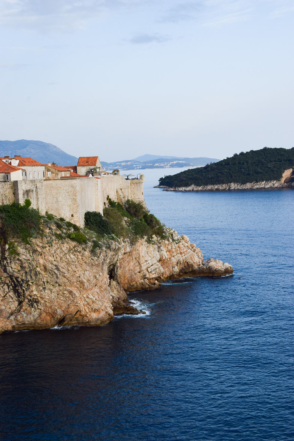 Green and rocky cliffs fall steeply to the sea with a small, stone city wall comes up from it with orange roofs and islands — travel photography by Shutter & Sojourn