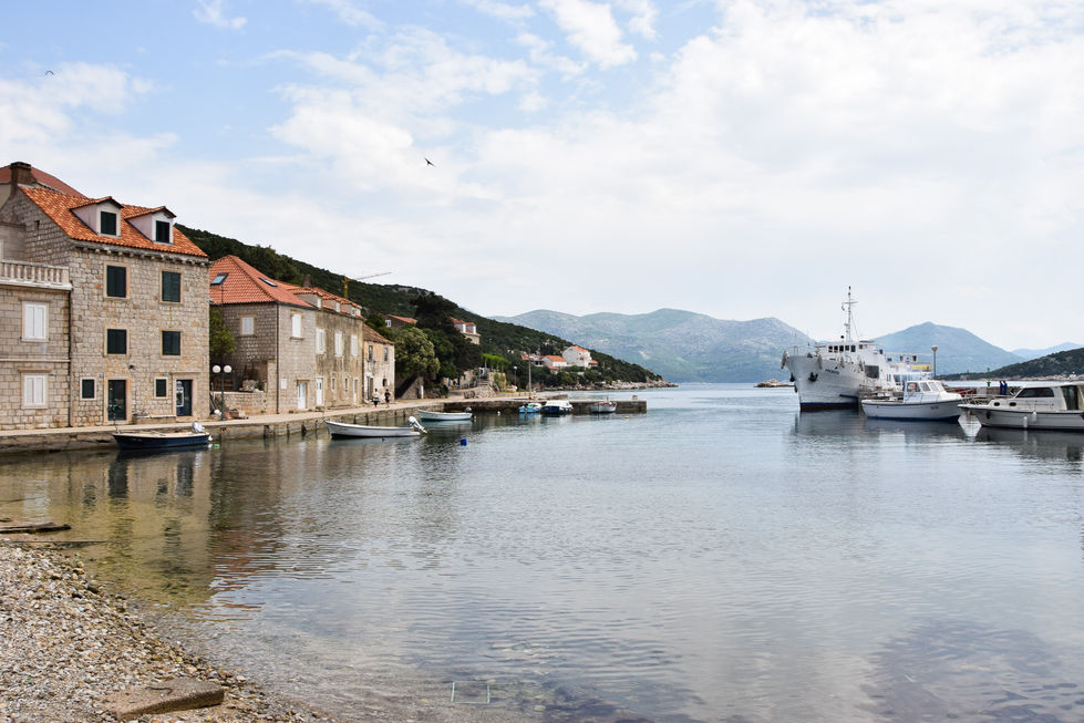 stone buildings with orange roofs line the water and marina with multiple boats tied up to the docks across the small bay. Mountains hazy in the background of Croatia — travel photography by Shutter & Sojourn