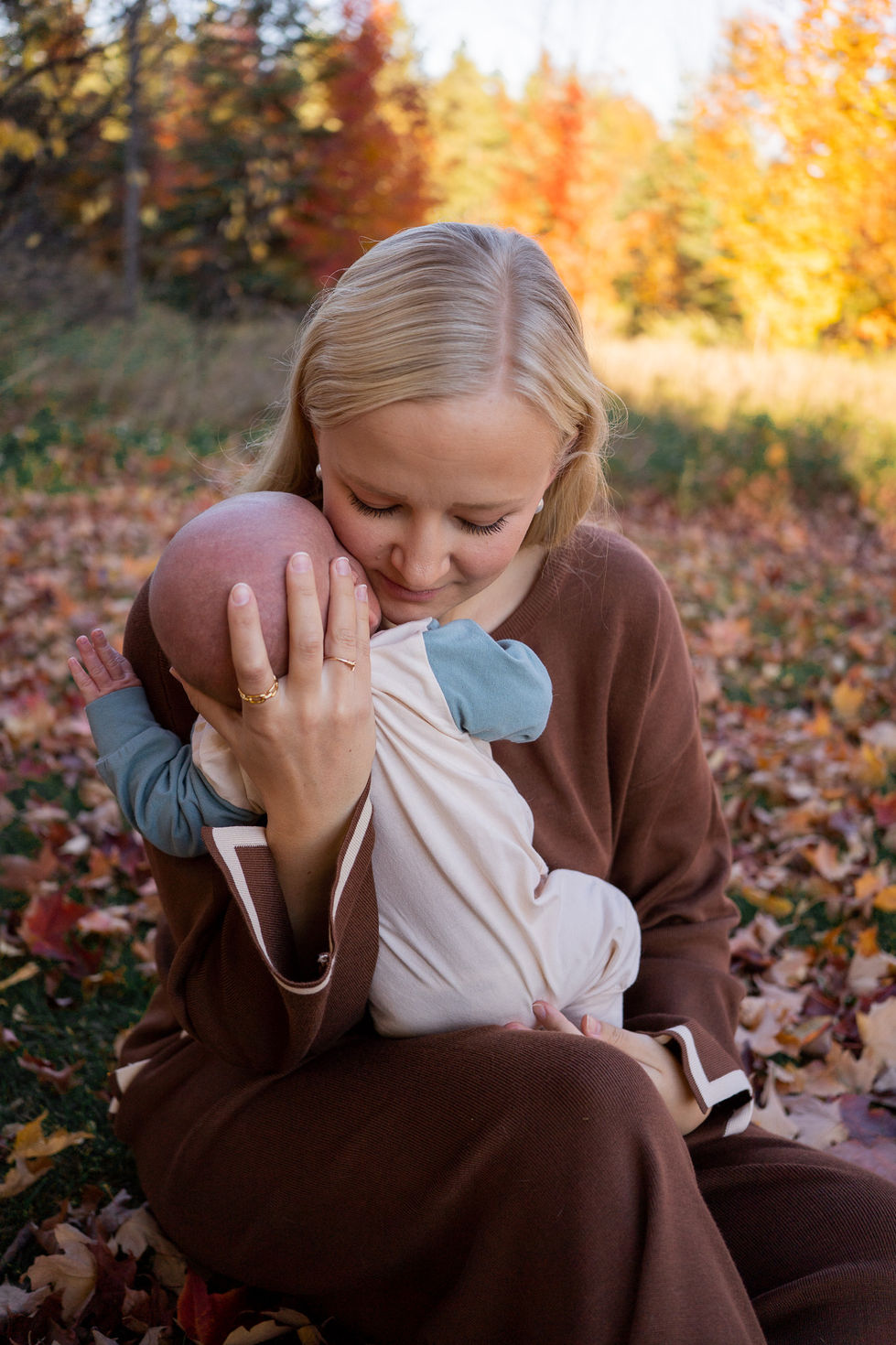 mother hugging newborn baby to her chest while wearing a brown matching set and sitting in a pile of fallen leaves — family photography by Shutter & Sojourn