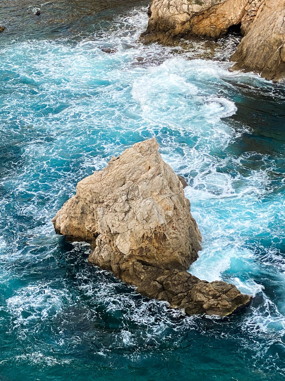 large boulder sticking out of a bright blue sea with white, foamy waves crashing around it — travel photography by Shutter & Sojourn