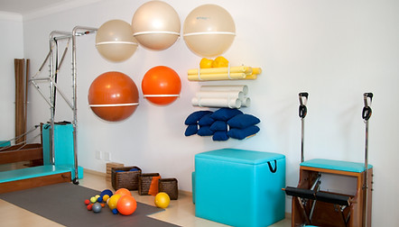 Outro ângulo da sala mostra a parede lateral. Ao fundo vemos parte dos aparelhos de pilates e as varas de madeiras apoiada na parede. Mais à frente, cinco estruturas de ferro que seguram as bolas maiores, sendo três delas brancas e duas laranjas. Abaixo delas vemos quatro cestos de palhas vazios e na sua frente várias bolas menores, de vários tamanhos e cores. Na parede também tem outras estruturas que guardam bolas menores, canos forrados e travesseiros pequenos que são usados no pescoço. Abaixo uma grande caixa forrada de verde, fechada. Ao lado outro aparelho de pilates, menor.