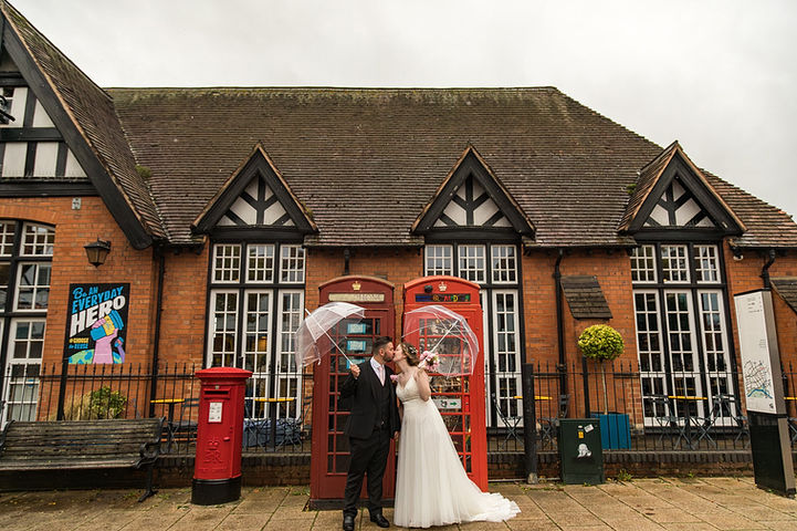 Couple pose by two red telephone boxes and a post box.