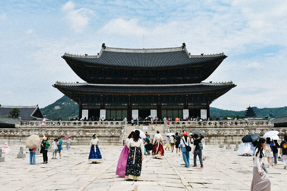 Visitors in colorful traditional outfits and umbrellas gather at Gyeongbokgung Palace, with its grand architecture under a blue, cloudy sky.