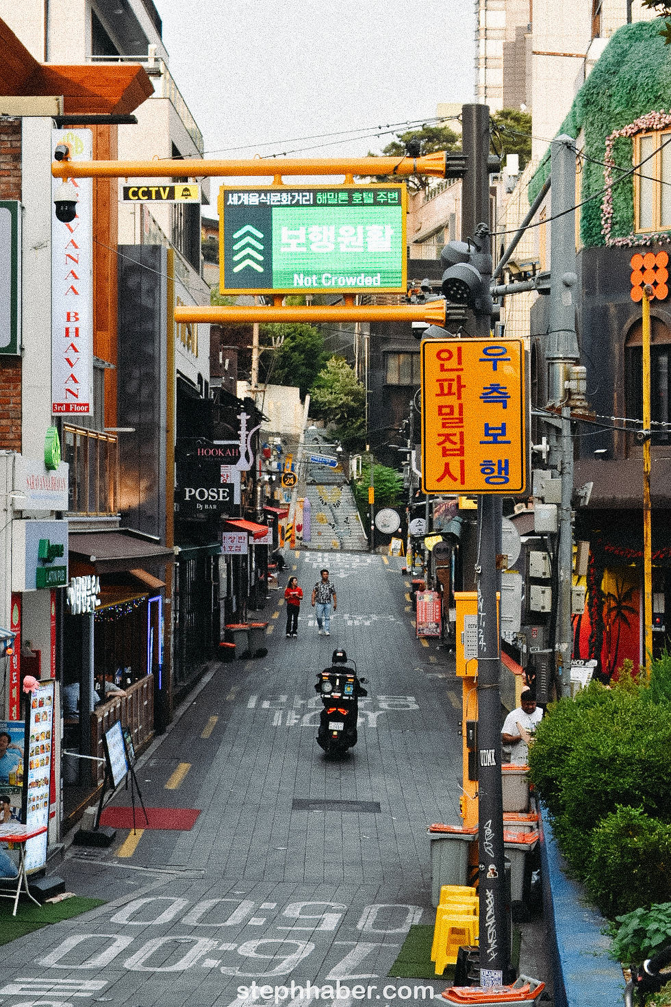 Busy urban street with people walking and a scooter riding. Colorful signs and buildings line the street. Sign reads "Not Crowded."