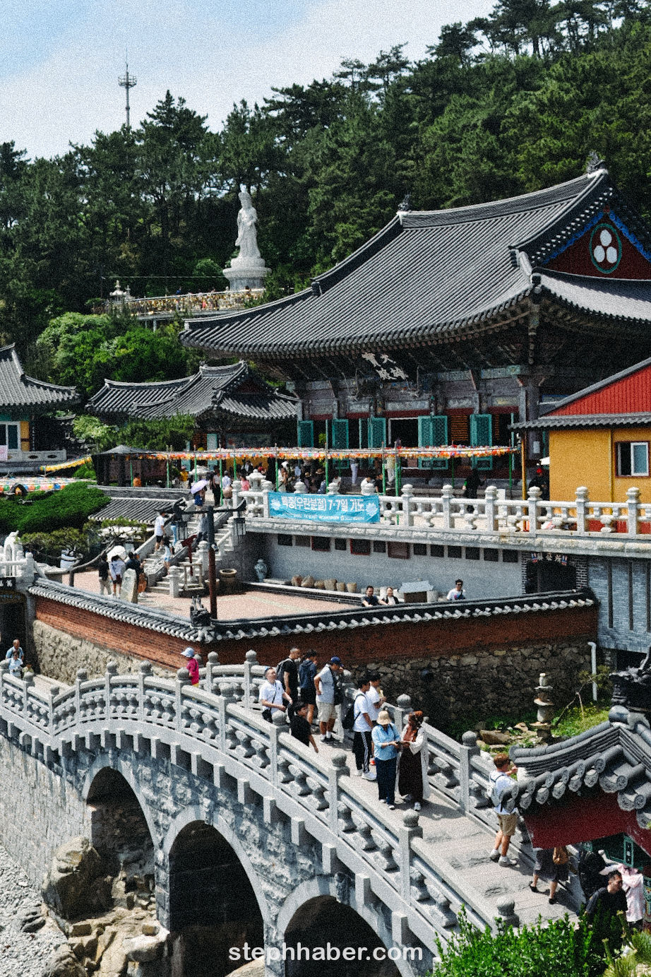 People walk on a stone bridge near traditional temple buildings. A large statue and trees are visible. Banner text and colorful lanterns adorn the scene.