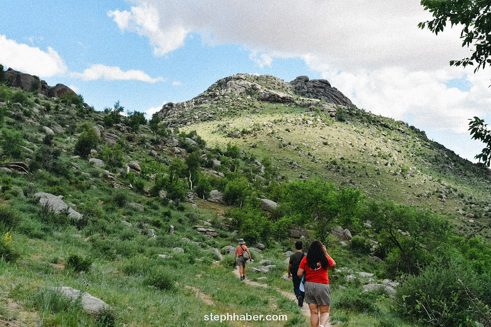 Three people hiking up a green, rocky hill under a blue, cloudy sky. One wears a red shirt. "stephhaber.com" visible at the bottom.