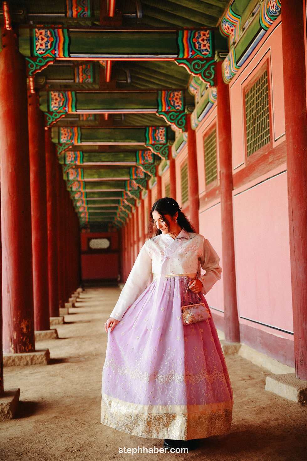 Woman in a lavender hanbok stands in a traditional Korean hallway with red columns and ornate patterns, looking down, exuding elegance.