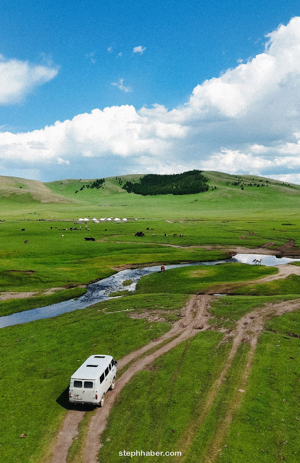 A white van travels on a dirt path through a lush green landscape with yurts, cows, and a stream under a bright blue sky.