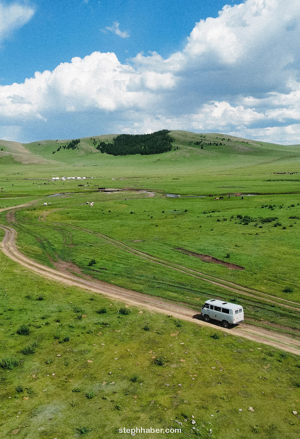 A white van drives on a dirt road through a vast green landscape under a blue sky with clouds. Hills and yurts are visible in the distance.