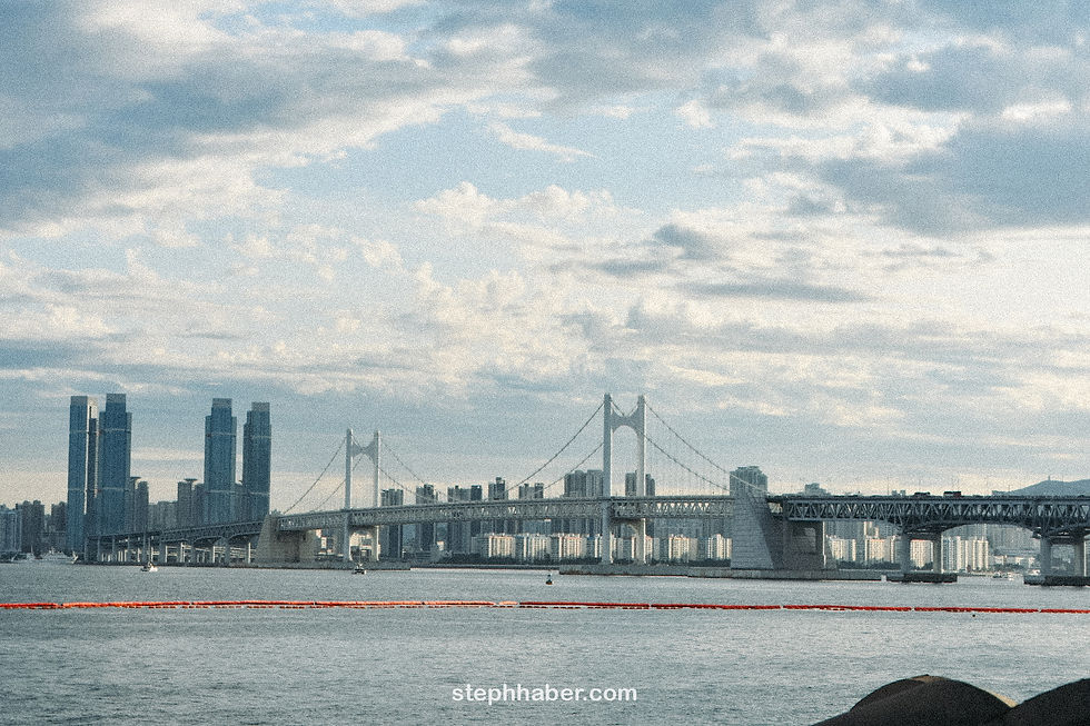Expansive view of a suspension bridge over water with tall city buildings in the background. Cloudy sky, red buoys in the foreground.