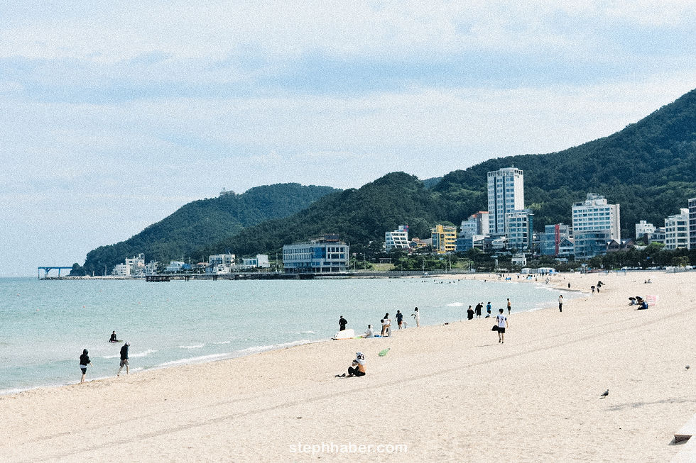 People on a sandy beach with a backdrop of tall, modern skyscrapers. Overcast sky sets a calm mood. Haeundae beach
