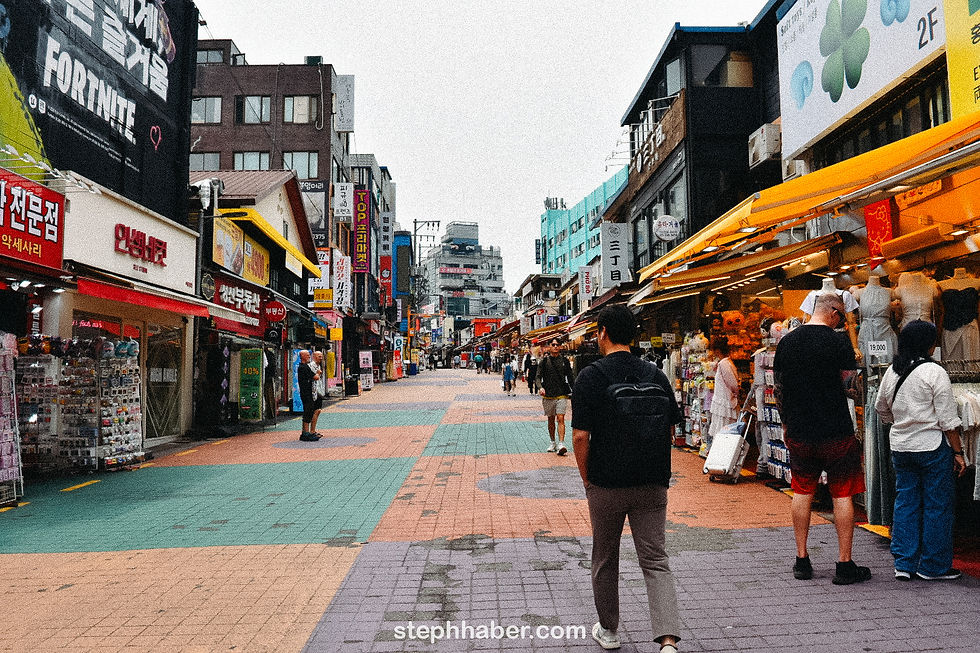 Busy street market with people walking and shopping. Colorful signs and awnings line the street. Overcast sky adds a muted tone.
