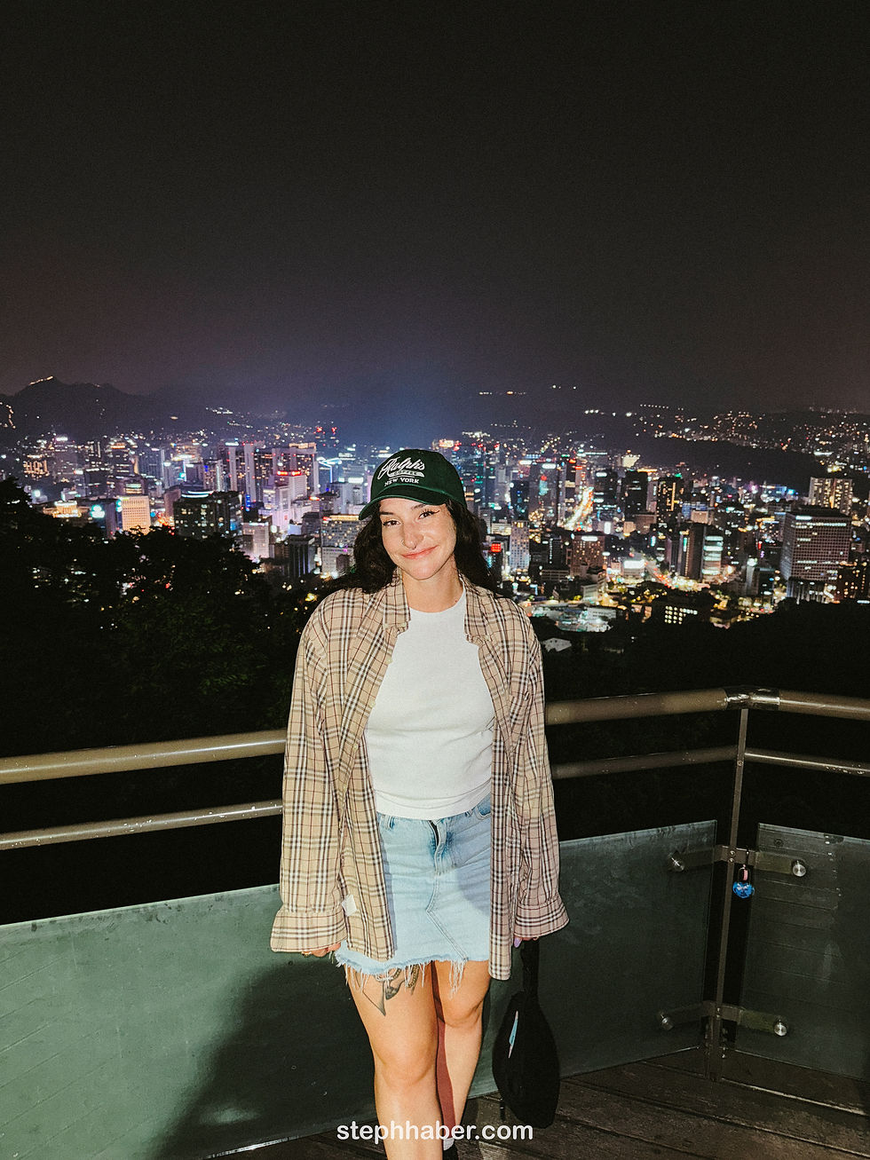 Woman in a plaid shirt and denim skirt smiles on a nighttime balcony overlooking a cityscape filled with lights. Text reads stephhaber.com.
