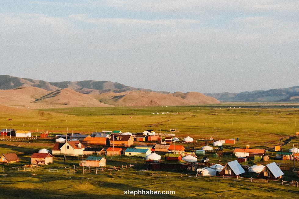 A village with colorful houses and yurts on a green plain, mountains in the background, under a clear sky. Visible text: stephhaber.com.