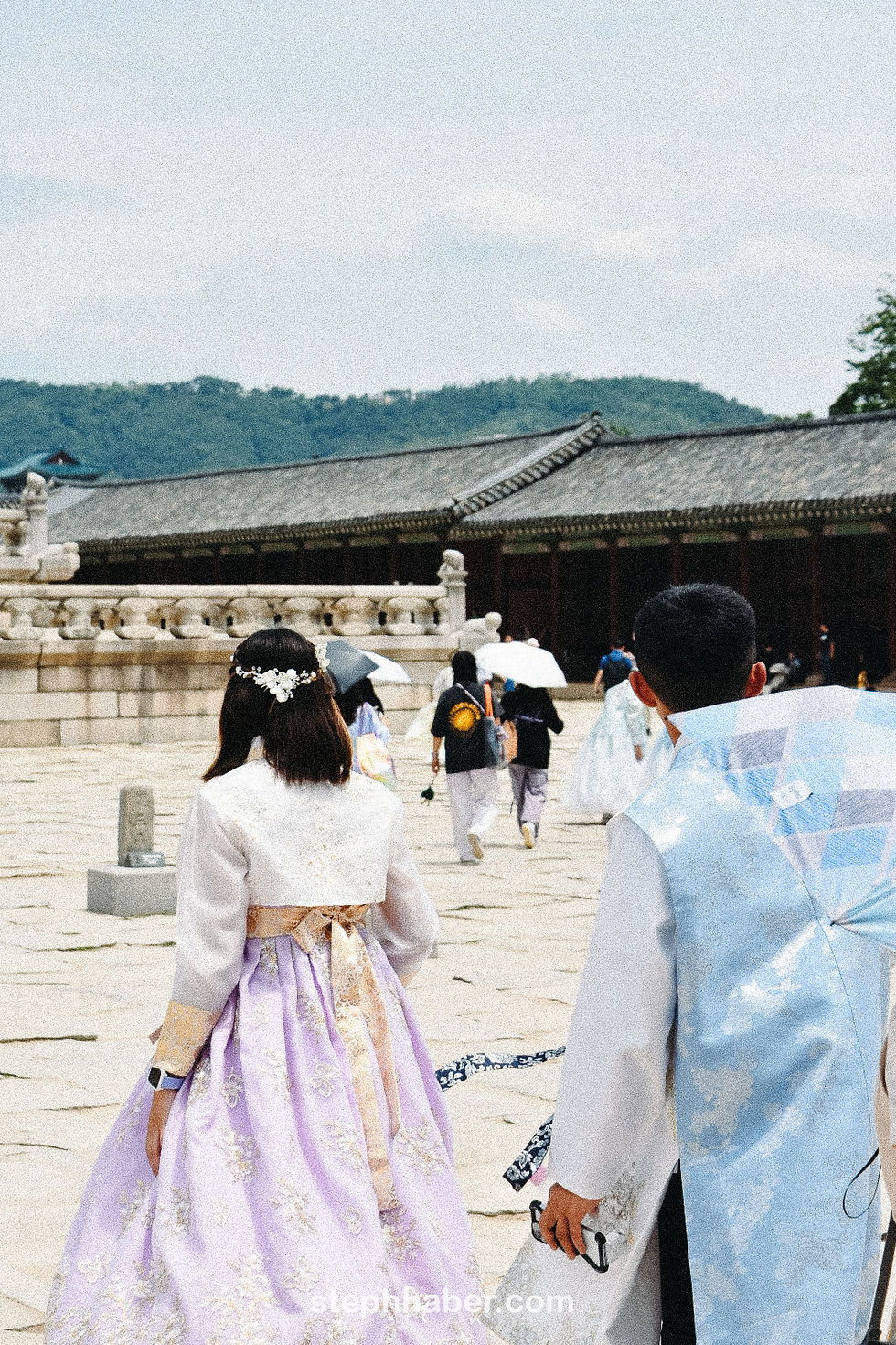 People in traditional Korean attire walk in a historical courtyard. One wears a lilac hanbok; others hold umbrellas. Background: hills, temple.