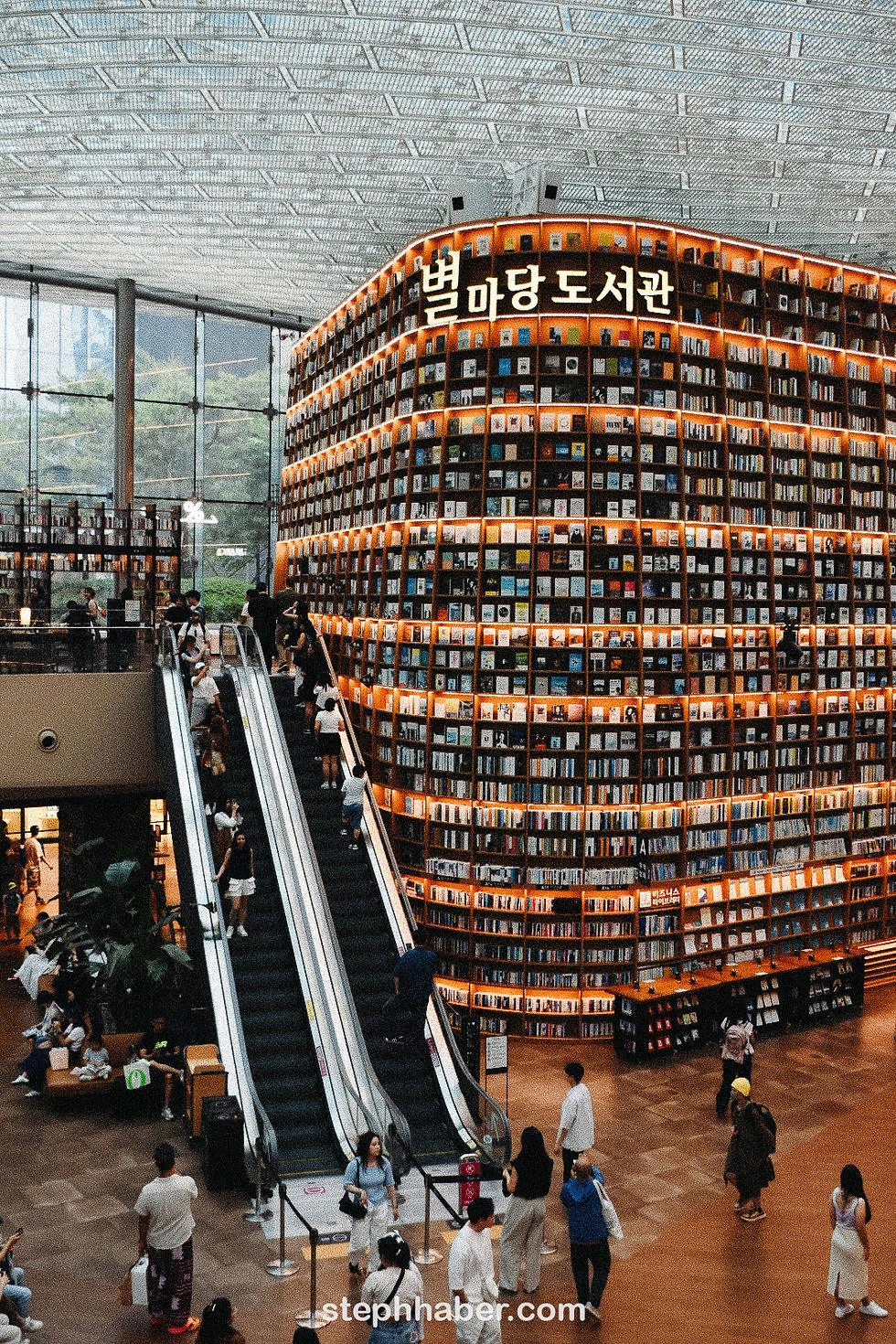 People on escalators in a spacious library with towering book displays, soft lighting, and a large ceiling. Text on signs in Korean.