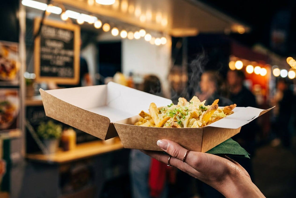A close-up shot of a street food customer holding an open kraft paperboard clamshell box filled with steaming loaded fries at an evening market. A blurred food truck with warm festoon lighting is visible in the background.