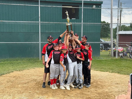 Youth baseball team in red-and-black Epsom jerseys huddles on a dirt field, lifting a trophy in celebration near a chain-link fence.