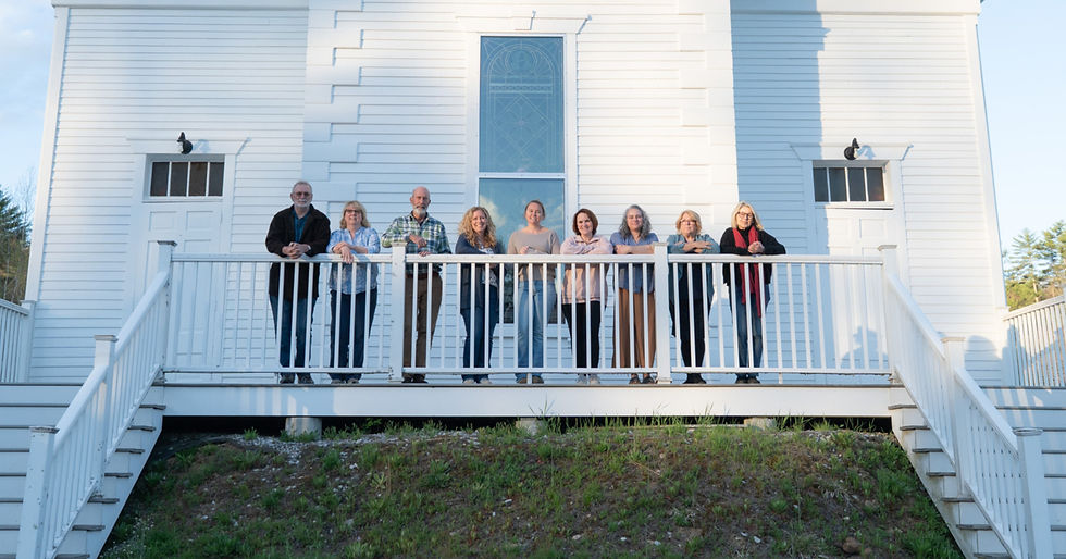 Members of the Epsom Tricentennial Committee lean on a white church porch railing, aka the Old Meeting House, in front of a tall window, smiling in bright afternoon light.