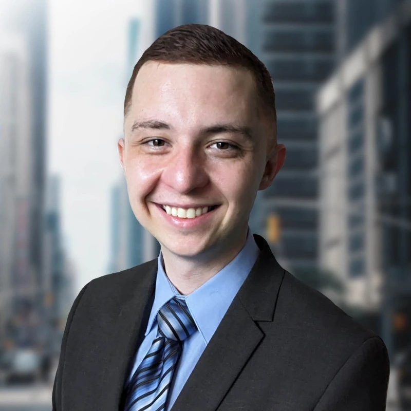 Brandon Brown smiling, in a dark suit, light blue shirt and striped tie, with city buildings behind him.